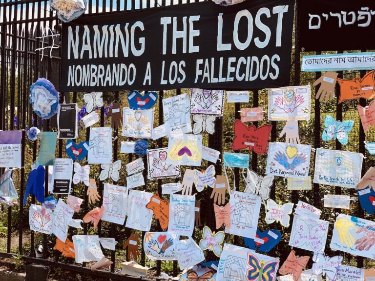 Photo of a black metal fence. Hanging from the fence are black signs with white lettering that say "Naming the Lost" in several languages. The fence is covered in brightly colored notes, many shaped like butterflies or hands. 